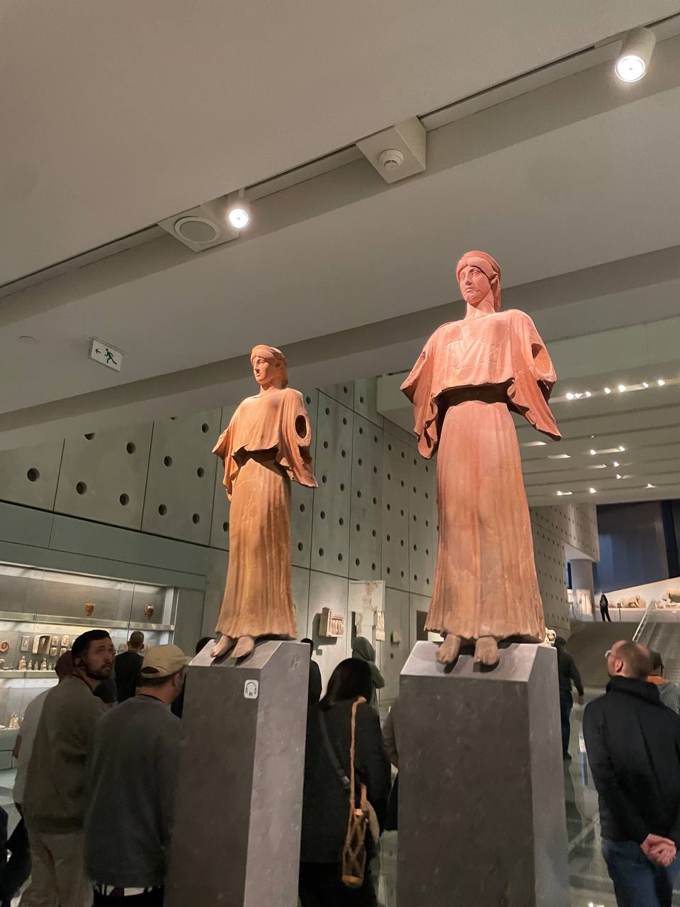 Two Nike statues in terracotta red at the Parthenon Museum, surrounded by visitors, in a large gallery space with display cases in the background