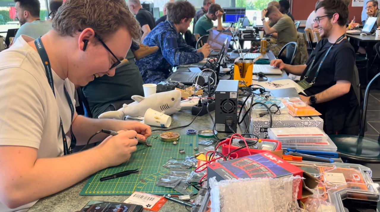 Participant soldering components at a hackathon workbench covered with electronic parts, cutting mat, multimeter, and a toy shark mascot, with more participants working in the background
