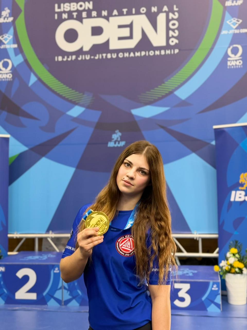Vladislava toont een gouden medaille voor het IBJJF Lisbon International Open 2026 banner, gekleed in het blauwe Brasa Belgium shirt