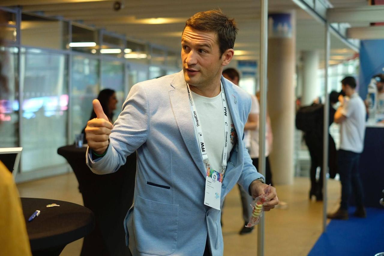 Frederik giving a thumbs up at the DrupalCon Prague booth in a light blue blazer, DrupalCon lanyard