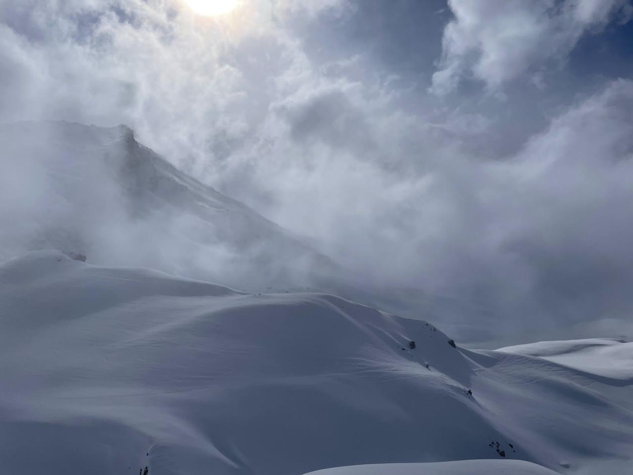 Alpine landscape with white snow dunes, peak barely visible through blowing clouds