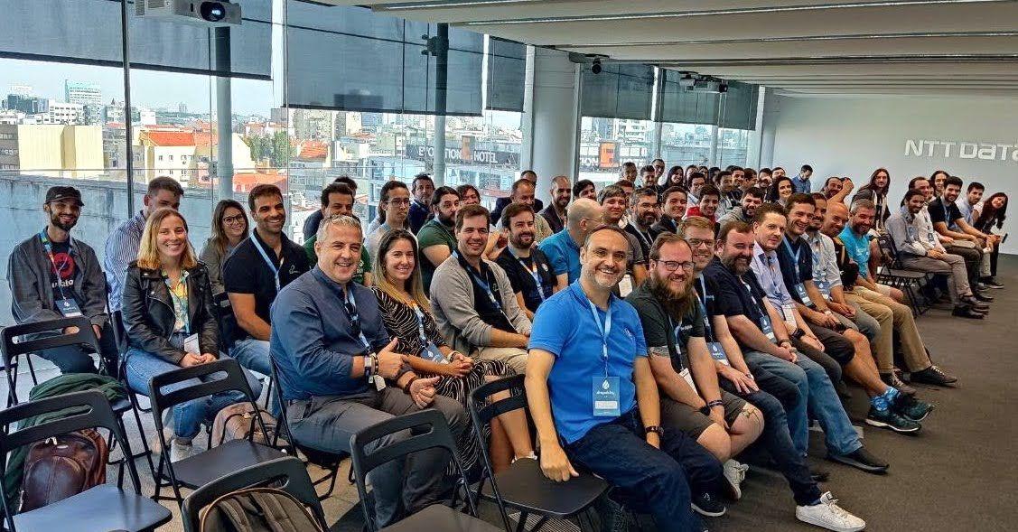 Drupal Day Portugal 2022 group photo at NTT DATA Atrium Saldanha - around 80 attendees in a bright conference room with Lisbon rooftops visible