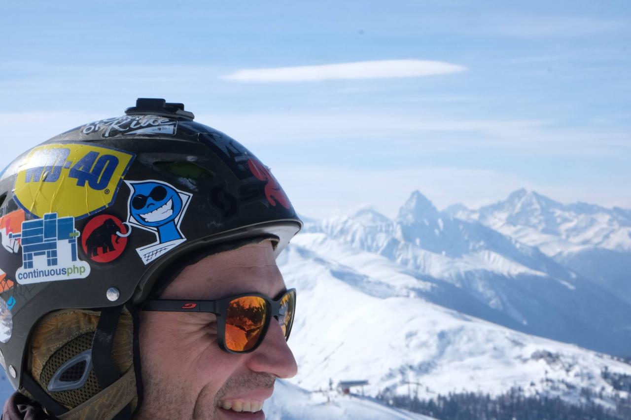 Skier at the summit with a sticker-covered helmet (WD-40, Drupal drop, ContinuousPHP) and panoramic alpine view