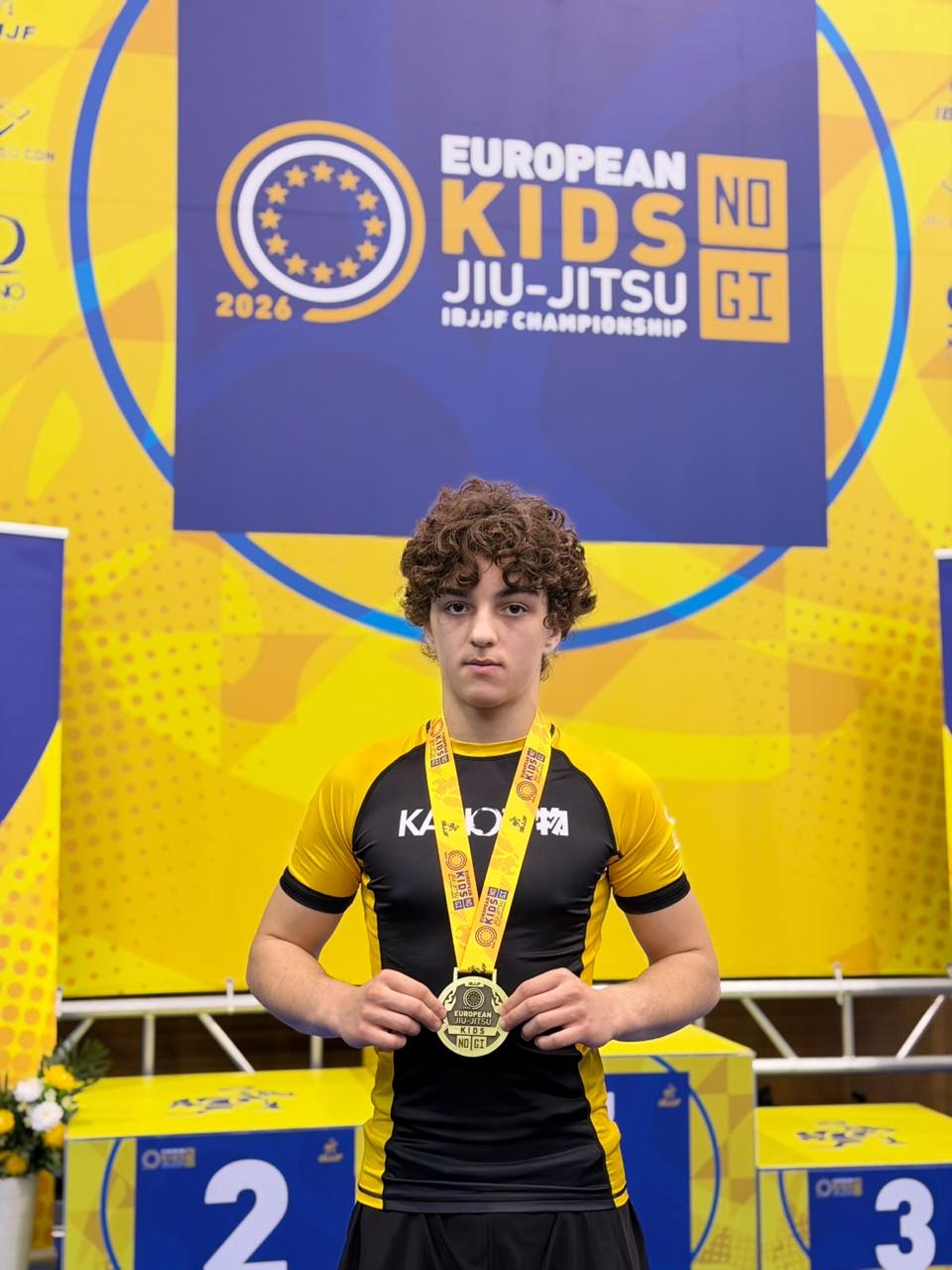 Ariyan met zijn bronzen medaille voor het IBJJF European Kids No-Gi Jiu-Jitsu Championship 2026 banner in Lissabon