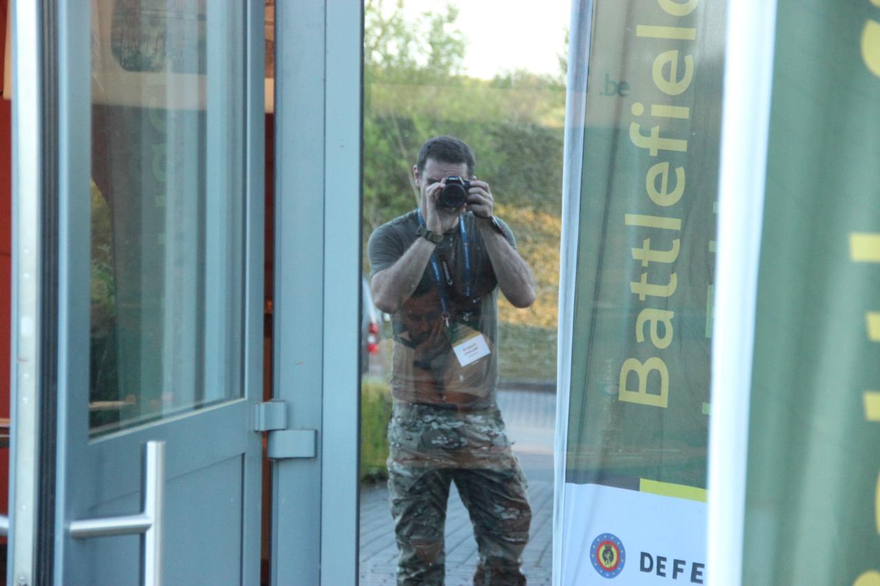 Frederik Wouters in military camouflage, photographing through a glass door reflection at the Battlefield of Things hackathon, with the event banner visible