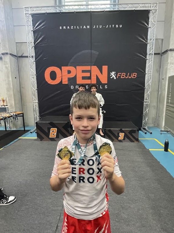 Alexander holding up two gold medals at the Brussels Open BJJ competition, in front of the FBJJB Open Brussels banner, wearing his Ferox rashguard