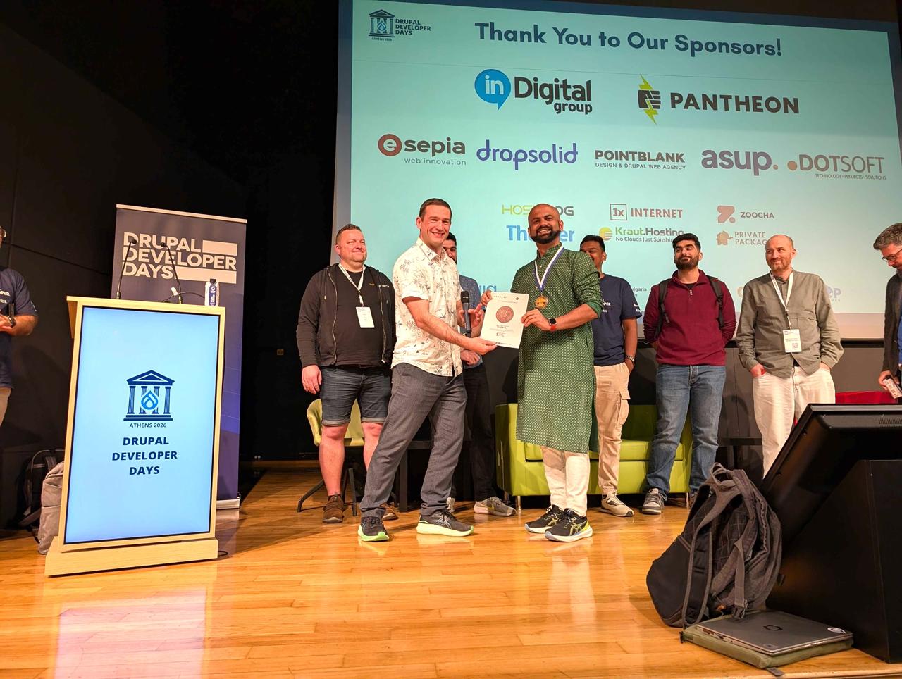 Anand receiving the bronze medal from Frederik Wouters on the main stage at Drupal Developer Days Athens 2026, large sponsor wall visible behind