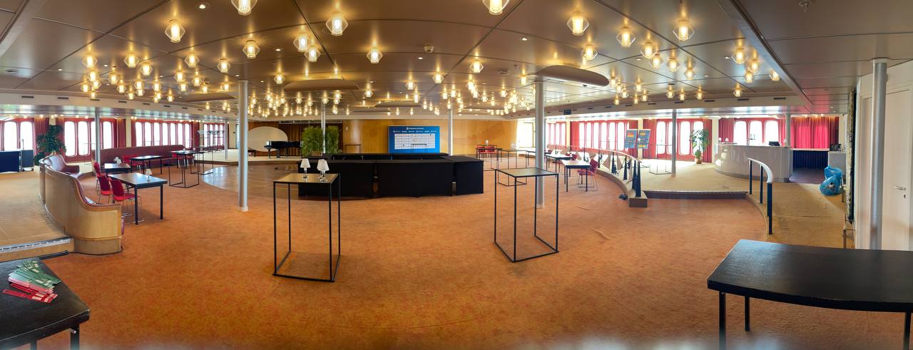 Panoramic view of the main hall on the SS Rotterdam, set up for the Enterprise AI Summit, with warm pendant lights across the ceiling and red-curtained portholes along the sides