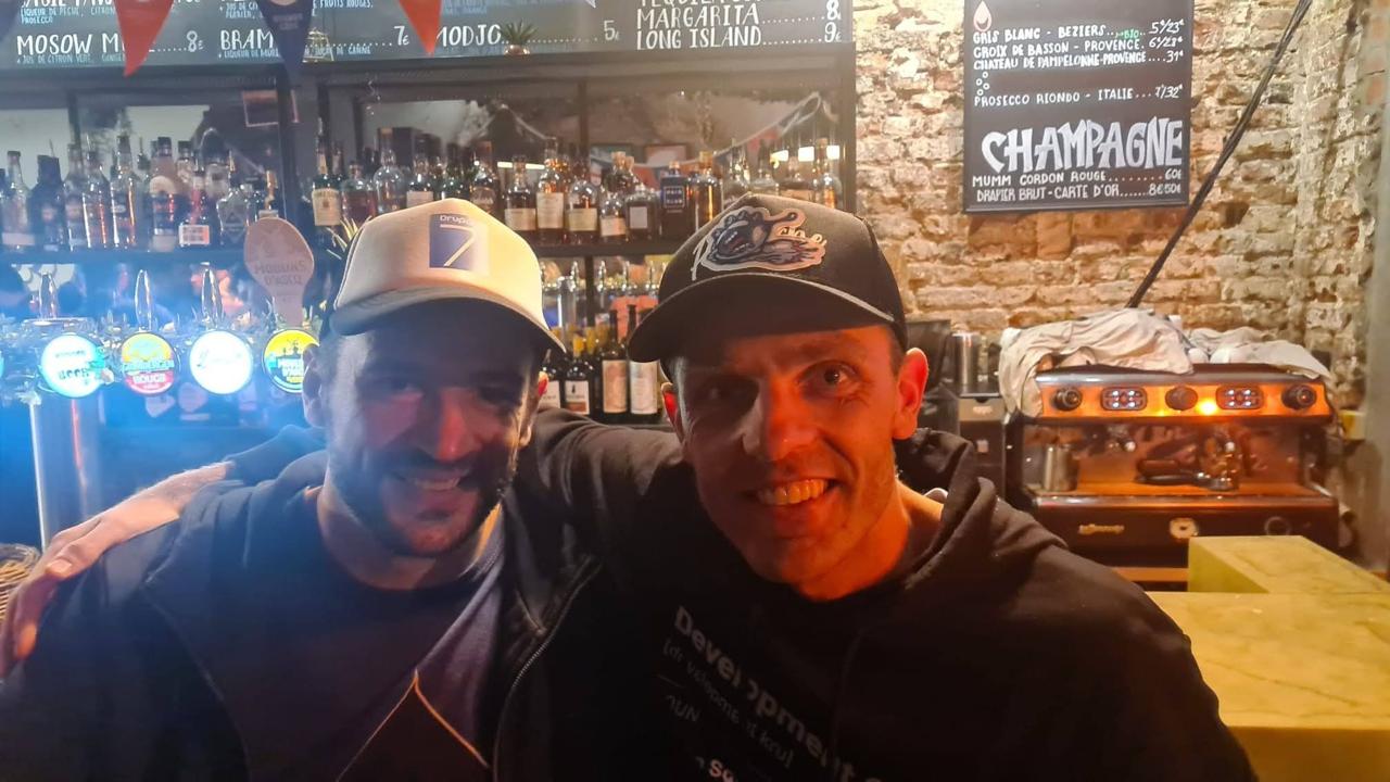 Two people in baseball caps at a Lille bar, smiling, one wearing a Development t-shirt
