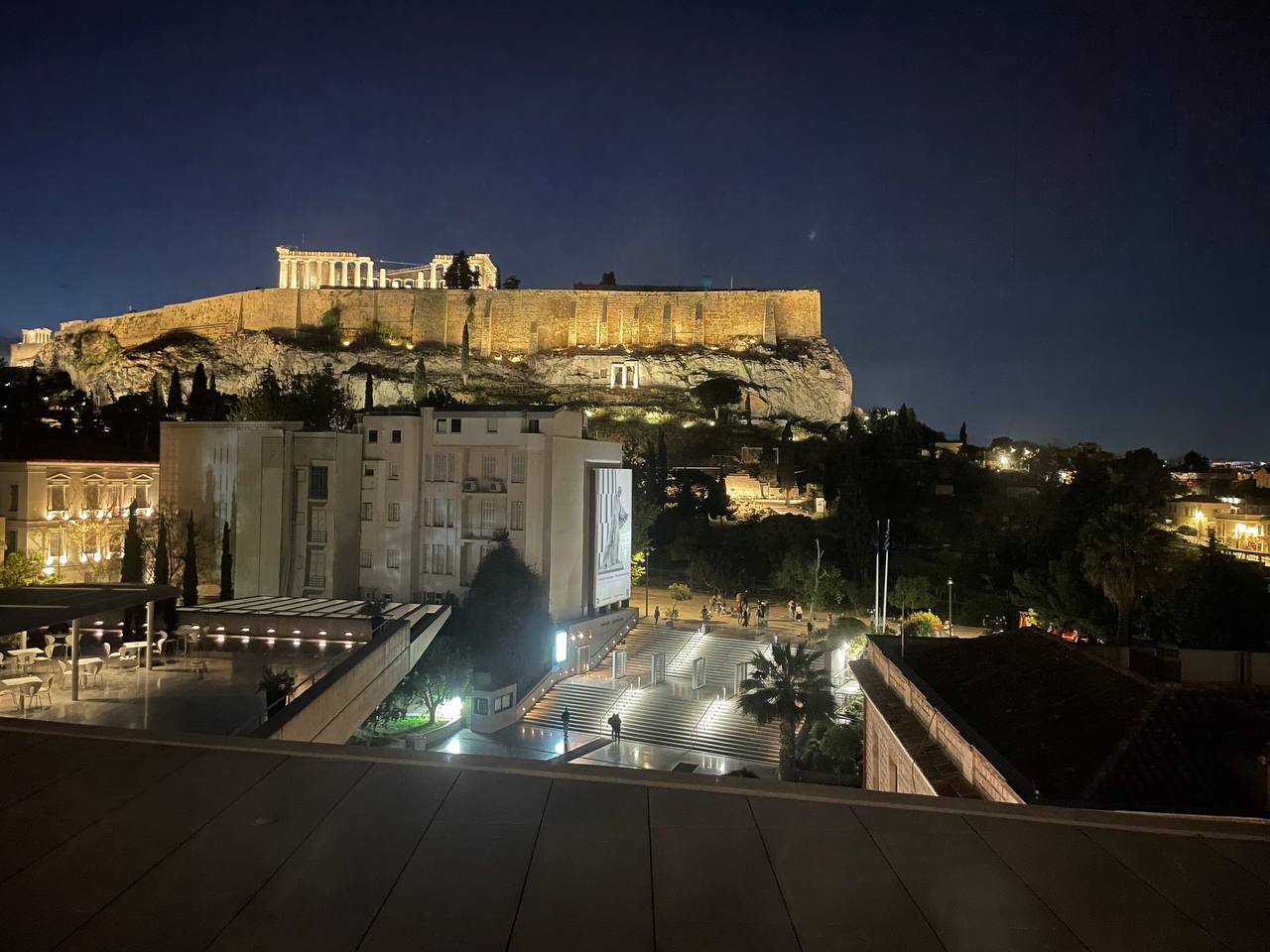 The Acropolis and Parthenon illuminated at night, viewed from the rooftop terrace of the Parthenon Museum, with the museum entrance and lit staircase in the foreground