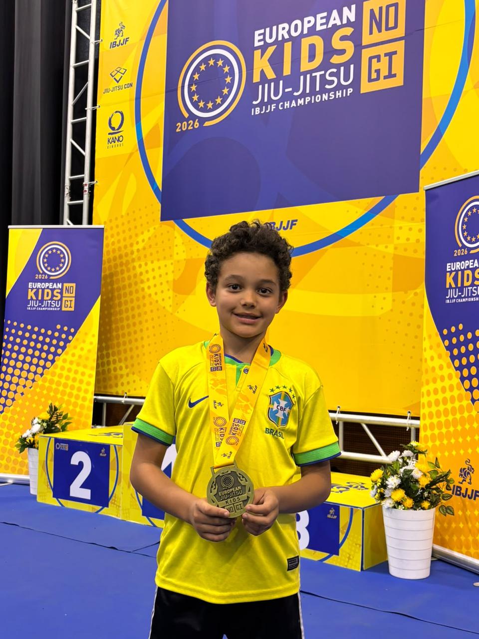 Adam in Braziliaans voetbalshirt voor het IBJJF European Kids No-Gi Jiu-Jitsu Championship 2026 banner, met zijn bronzen medaille om de nek, naast de tweede podiumplek