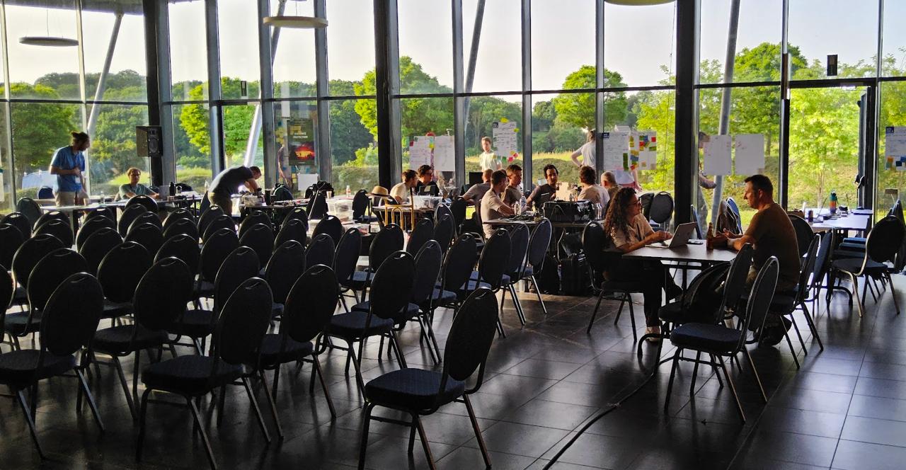 Hackathon workspace overview with participants at tables working on laptops, post-its on the walls, and military and civilian participants mixing freely