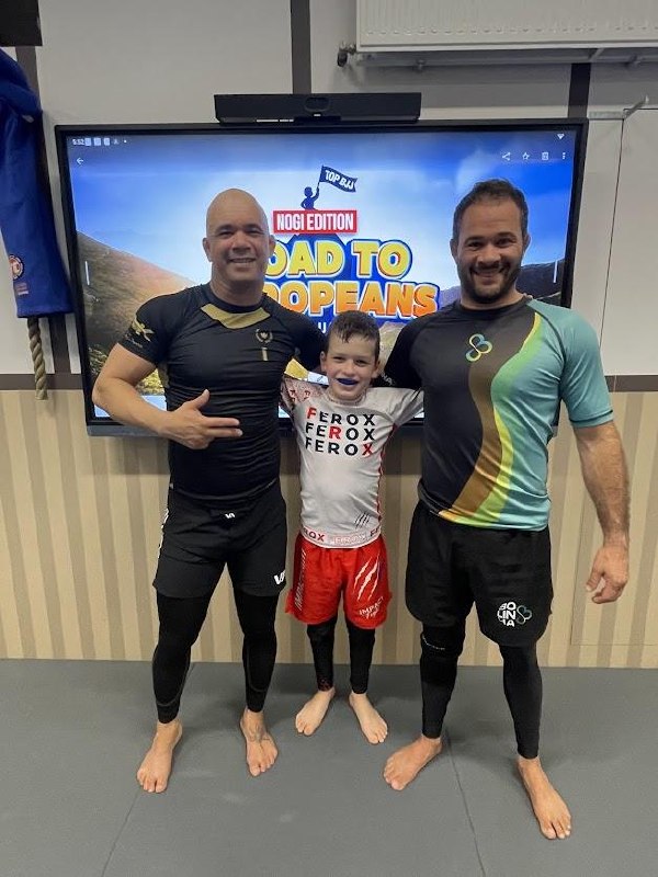 Alexander standing between two adult BJJ coaches in front of a Road to Europeans Nogi Edition screen at the Tilburg gym, all three smiling