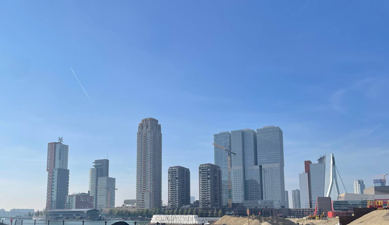 Rotterdam skyline on a clear blue-sky day, with the Erasmus Bridge visible on the right and high-rise towers along the Maas riverbank