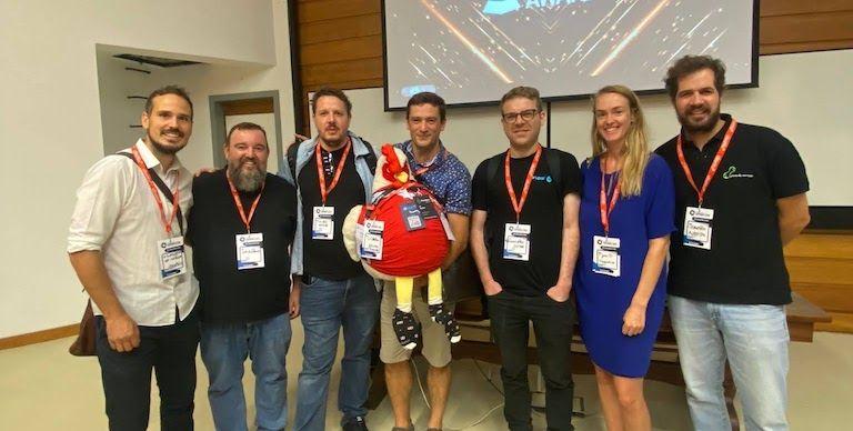 Group photo at Drupal Day Portugal 2023 - seven people with Lil Rooster mascot in front of the event stage