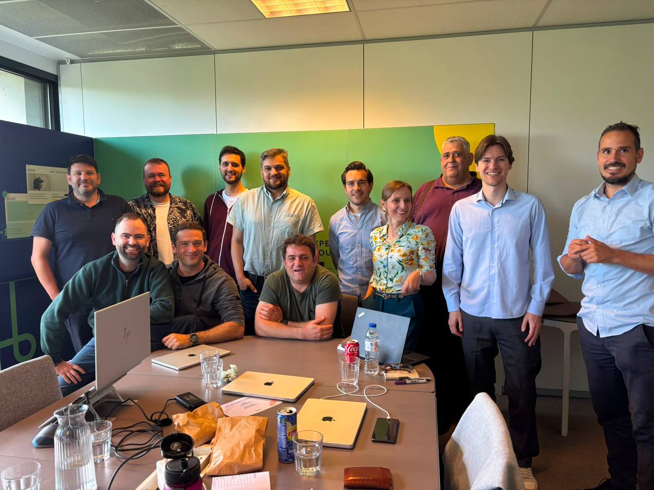 The Dropsolid AI management team gathered in a conference room with green and yellow wall panels and the Dropsolid logo, a mix of people sitting and standing around a table with laptops