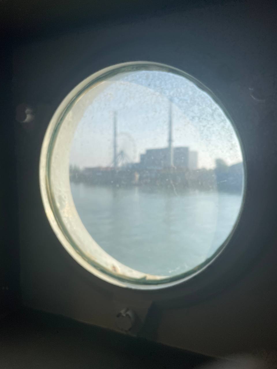 View through a porthole window on the SS Rotterdam, looking out over the Maas with the Rotterdam industrial skyline in the background