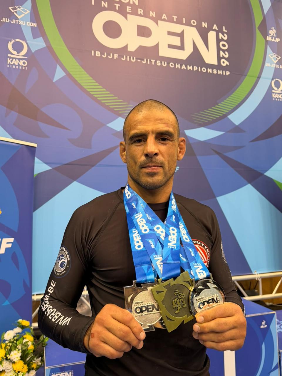 Coach Wim Deputter toont drie medailles (zilver, goud en brons) voor het IBJJF Lisbon International Open 2026 banner, gekleed in een zwart rashguard van Brasa Belgium