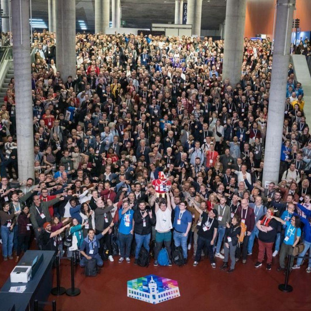 Aerial group photo of 1300 DrupalCon Lille attendees filling the Grand Palais atrium, someone holding up Lil Rooster in the centre