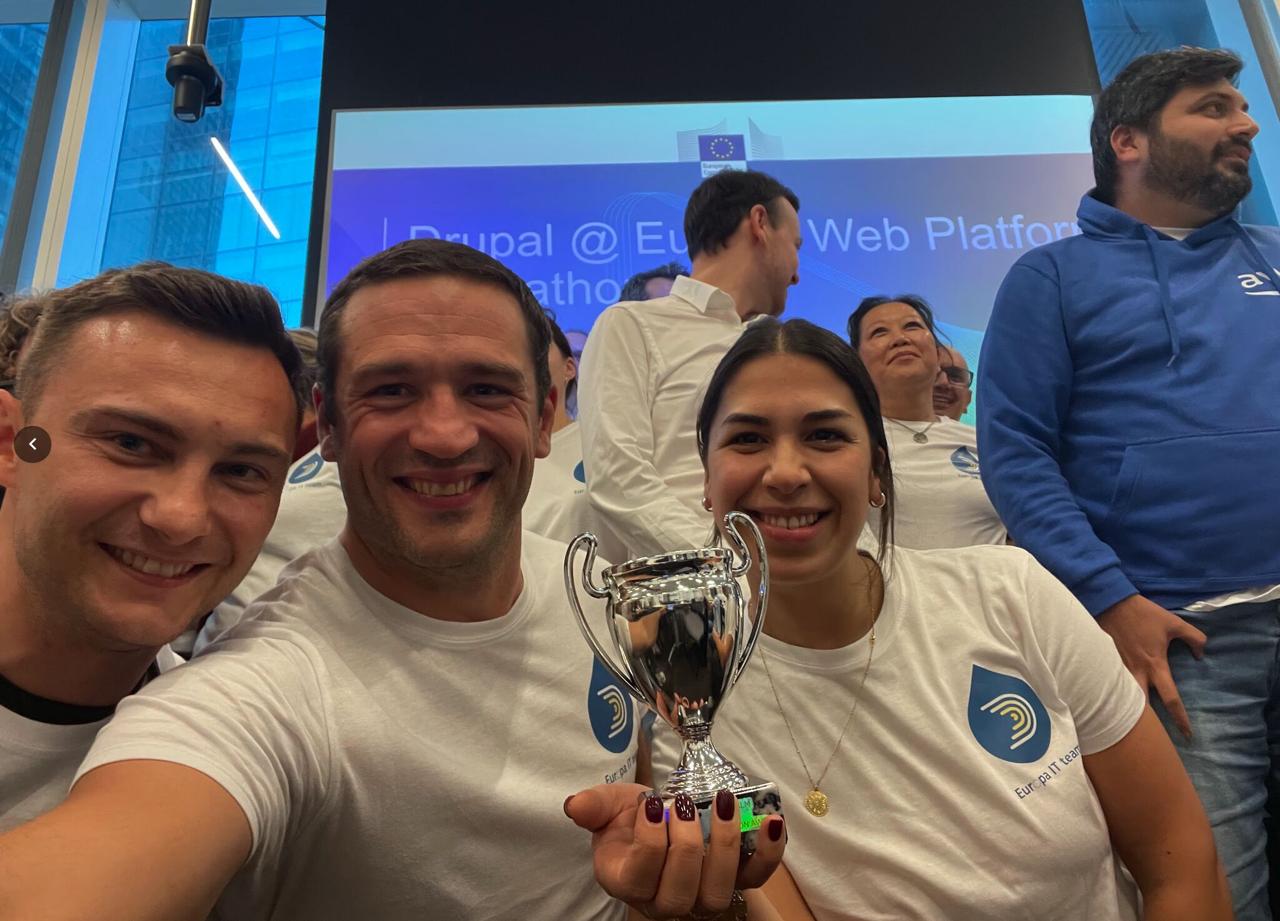 Three Squad 4 members posing with the silver cup trophy, Drupal Europa Web Platform Hackathon banner in background
