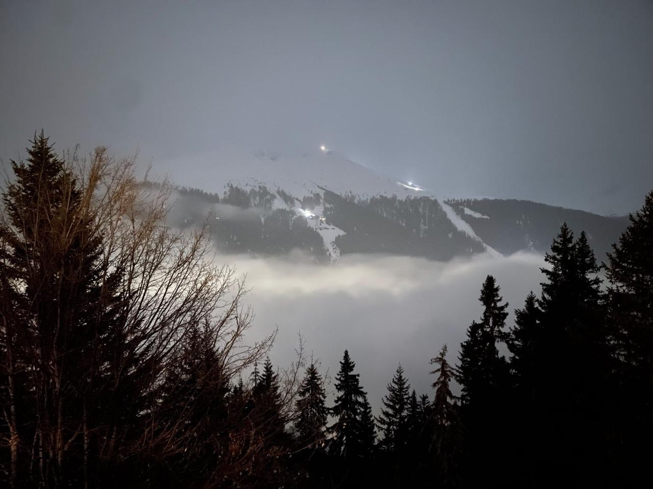 Nebelmeer at night: ski slope lights visible above a sea of fog, silhouetted trees in foreground