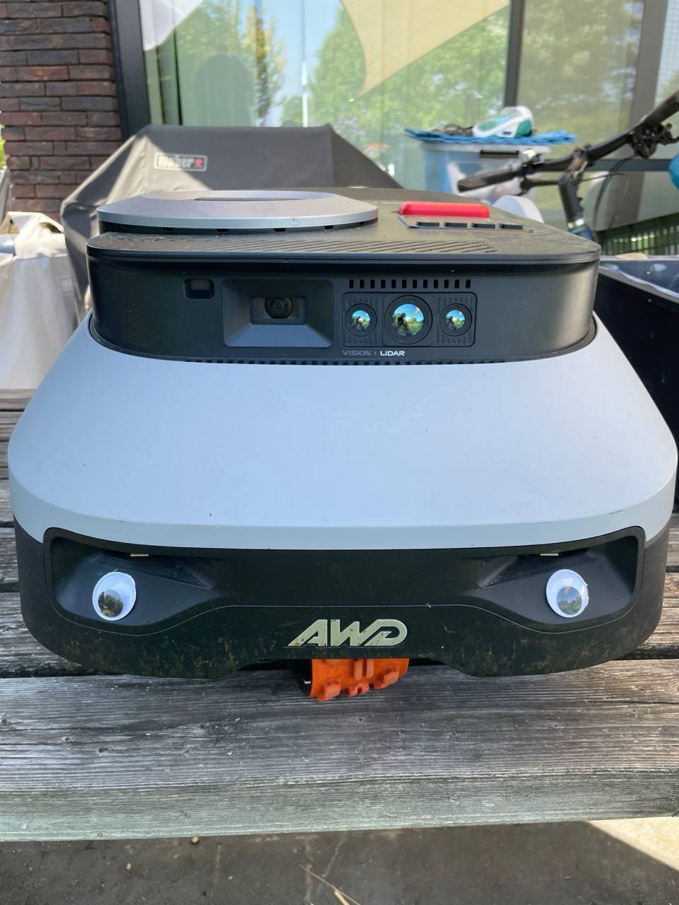 Segway Navimow i220 AWD robotic mower with two googly eyes stuck to the front, sitting on a garden terrace, LiDAR and vision sensors visible on top