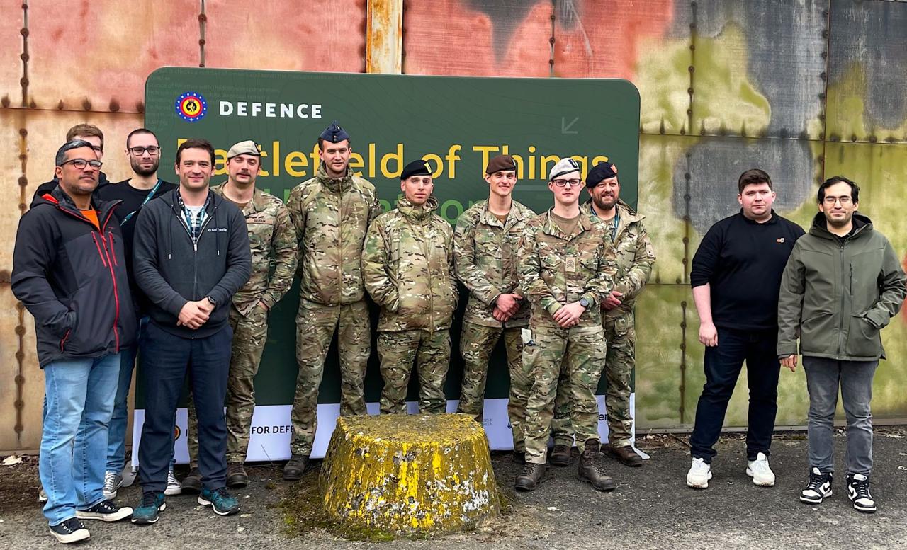 Mixed team of civilians and Belgian Army soldiers posing together in front of the Battlefield of Things sign outside