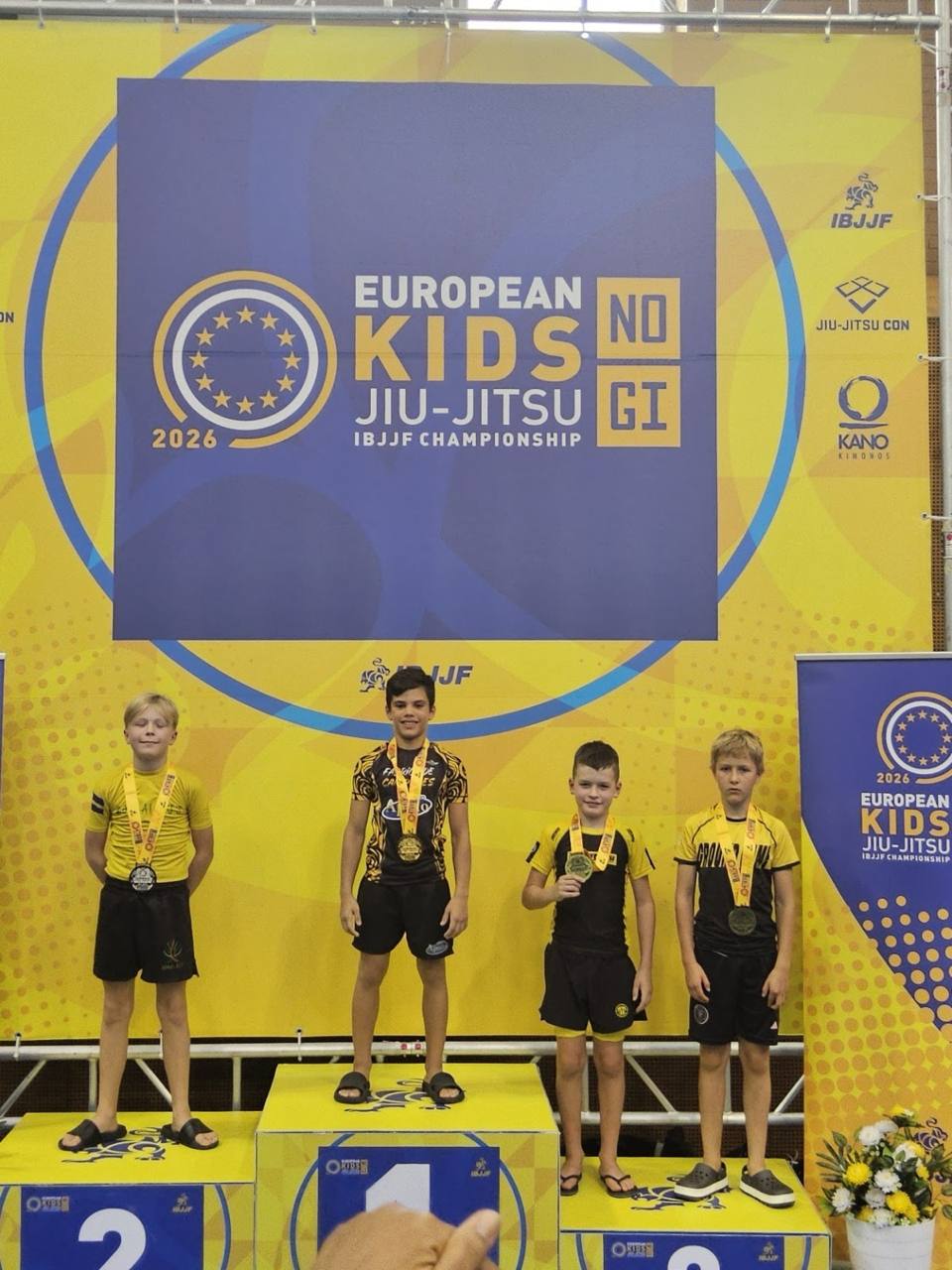 Alexander standing on the number 1 podium spot at the IBJJF European Kids No-Gi Jiu-Jitsu Championship 2026, wearing his gold medal, surrounded by three other competitors on the podium in front of the yellow and blue IBJJF banner
