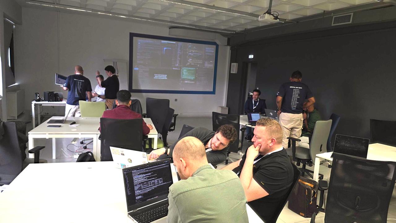 Overview of the Vibe Coding Challenge room: four competitors at their laptops with the live OBS screen stream projected on the wall behind them