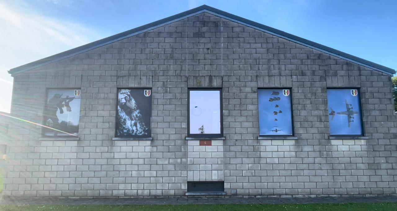 A grey cinder block building at the Belgian Commando Training Center in Marche-les-Dames, with four windows decorated with Belgian Defence imagery: commandos rappelling, paratroopers jumping, and a military transport aircraft