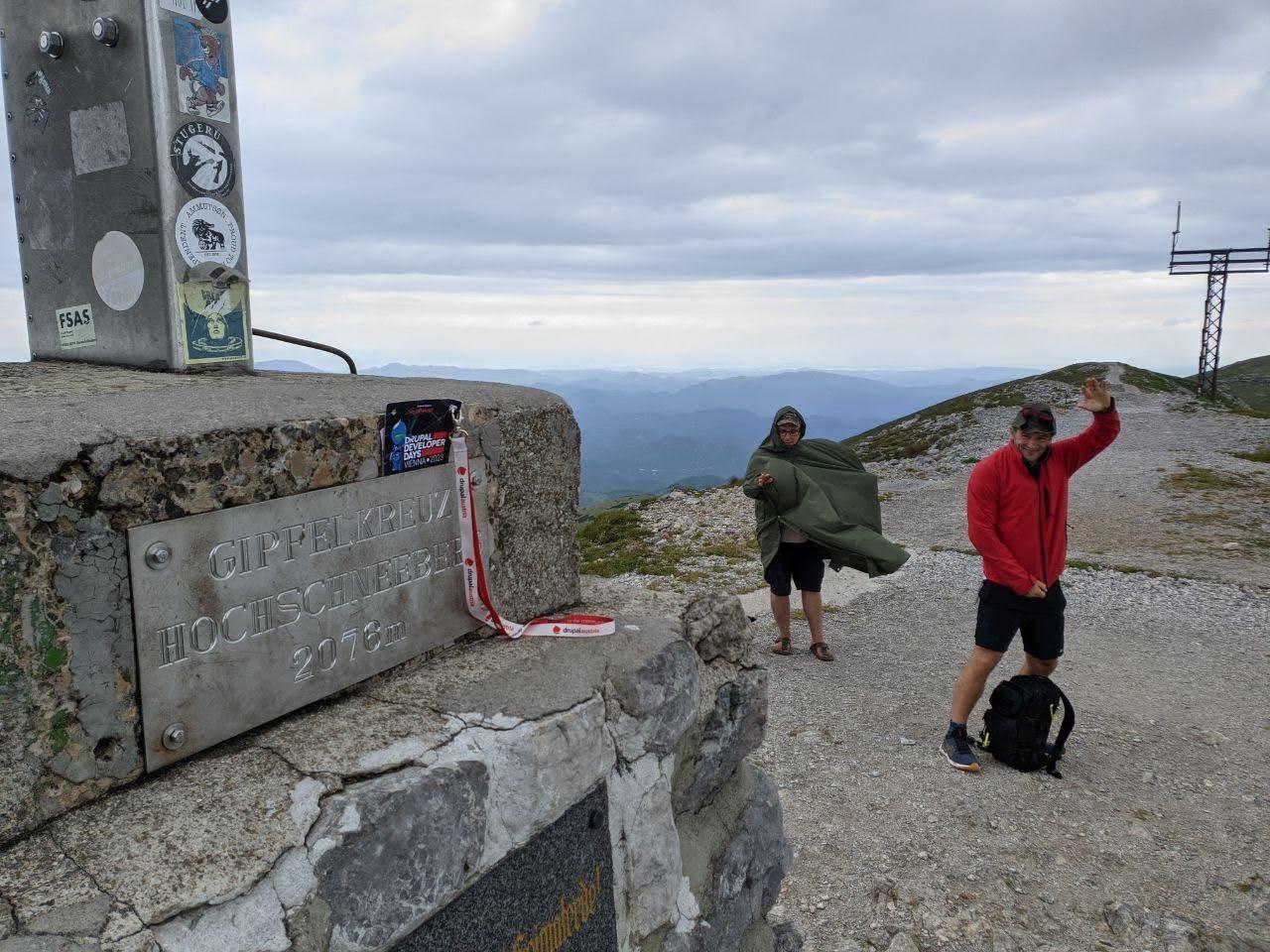 Two Drupal hikers at Gipfelkreuz Hochschneeberg 2076m - one wrapped in an army blanket, Frederik in red jacket waving, Drupal Developer Days Vienna 2023 lanyard on the cross
