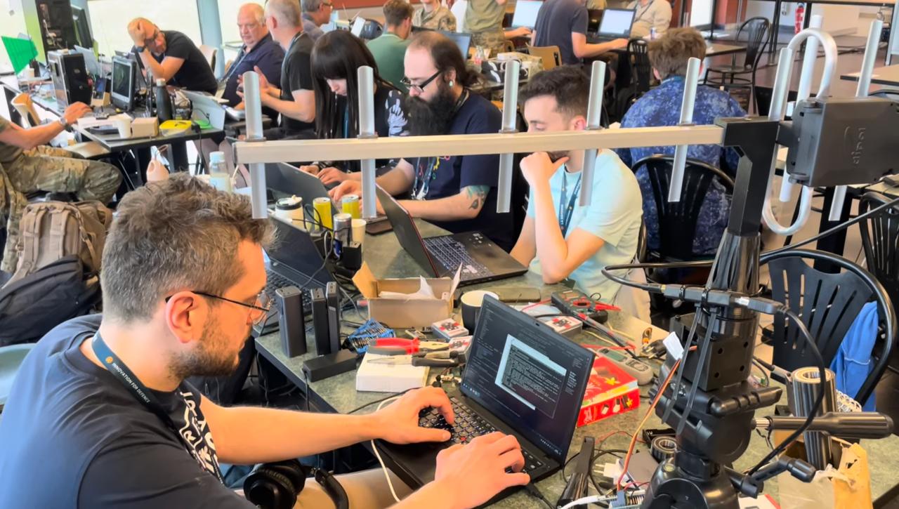 Close-up of hackathon participants at a crowded workbench covered in laptops, tools, cables, and electronic components, with antenna mounts visible and a terminal window open on screen